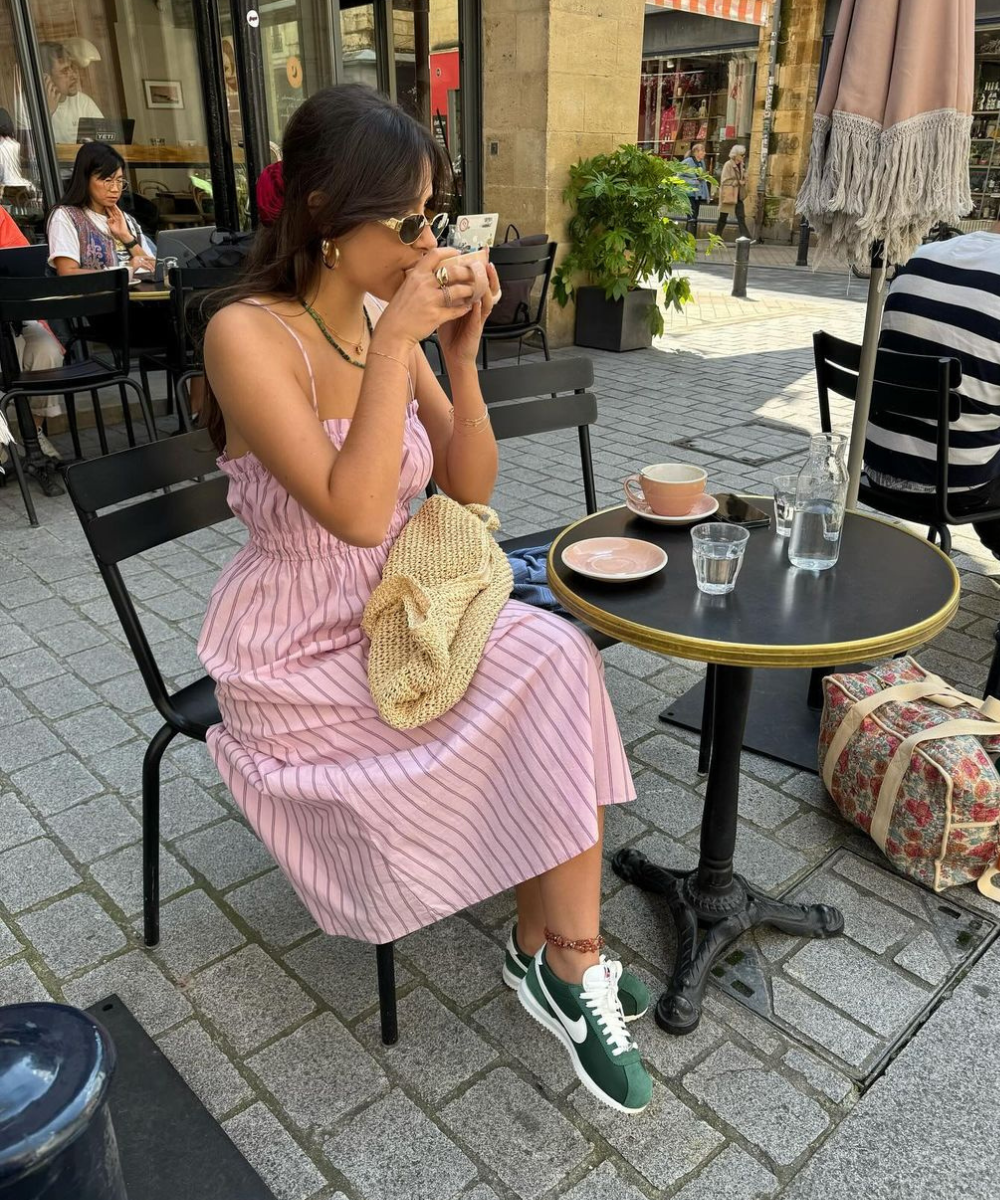 Mulher em vestido rosa listrado, óculos de sol, tênis verde e bolsa de crochê sentada em café ao ar livre. Estilo casual e descontraído, combinando tendências de moda sustentável e confortável.