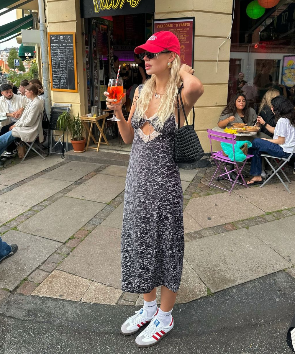 Mulher vestindo vestido midi estampado, com tênis Adidas, boné vermelho e óculos de sol. Ela segura um drink e carrega bolsa de crochê. Estilo casual-chique, tendência streetwear com mistura de peças esportivas e elegantes. Ambiente de café ao ar livre.