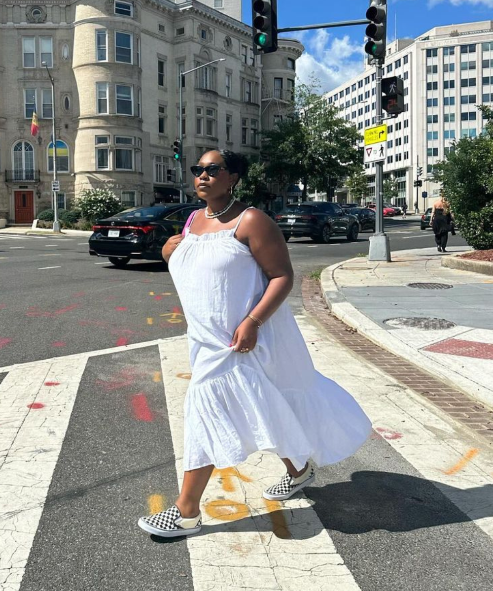 Mulher caminhando na rua com vestido branco longo e esvoaçante, óculos de sol estilo retrô e tênis xadrez preto e branco. Look casual e confortável, destacando tendências de moda urbana e acessível.