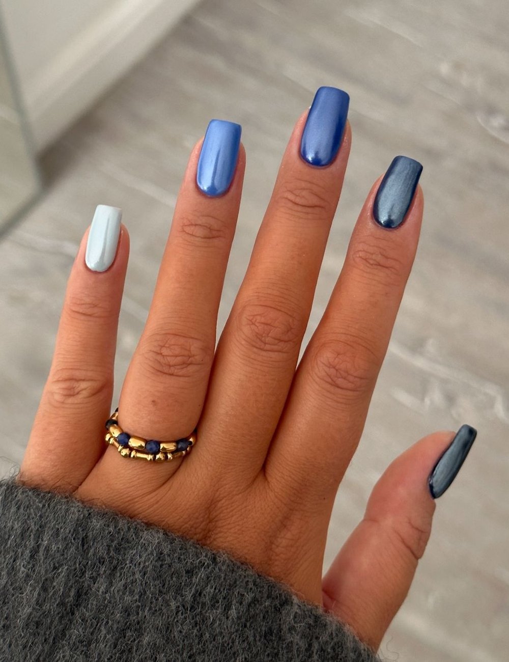 Unhas com cor de esmalte azul, com acabamento brilhante, seguindo a tendência de nail art sofisticada. Anel dourado com detalhes em pedras azuis e mangas de blusa cinza, sugerindo um look moderno e elegante de outono/inverno.