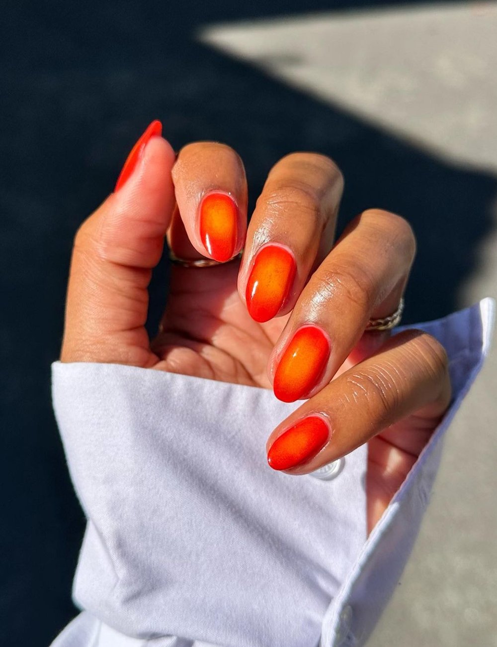 Unhas com  cor de esmalte em degradê laranja-avermelhado, tendência vibrante nas manicures modernas. A mão está vestida com uma elegante camisa branca de manga longa, destacando um visual sofisticado e contemporâneo.