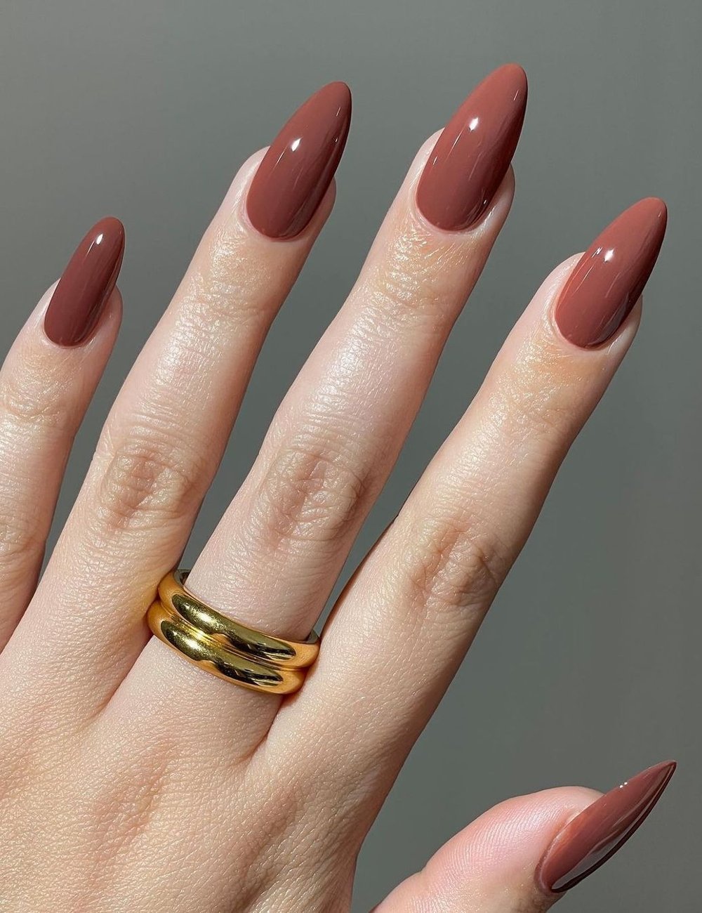 Unhas alongadas em formato amendoado, pintadas com cor de esmalte marrom brilhante. A mão exibe um anel duplo dourado, destacando uma combinação elegante e moderna. A tendência reflete sofisticação e minimalismo na moda.