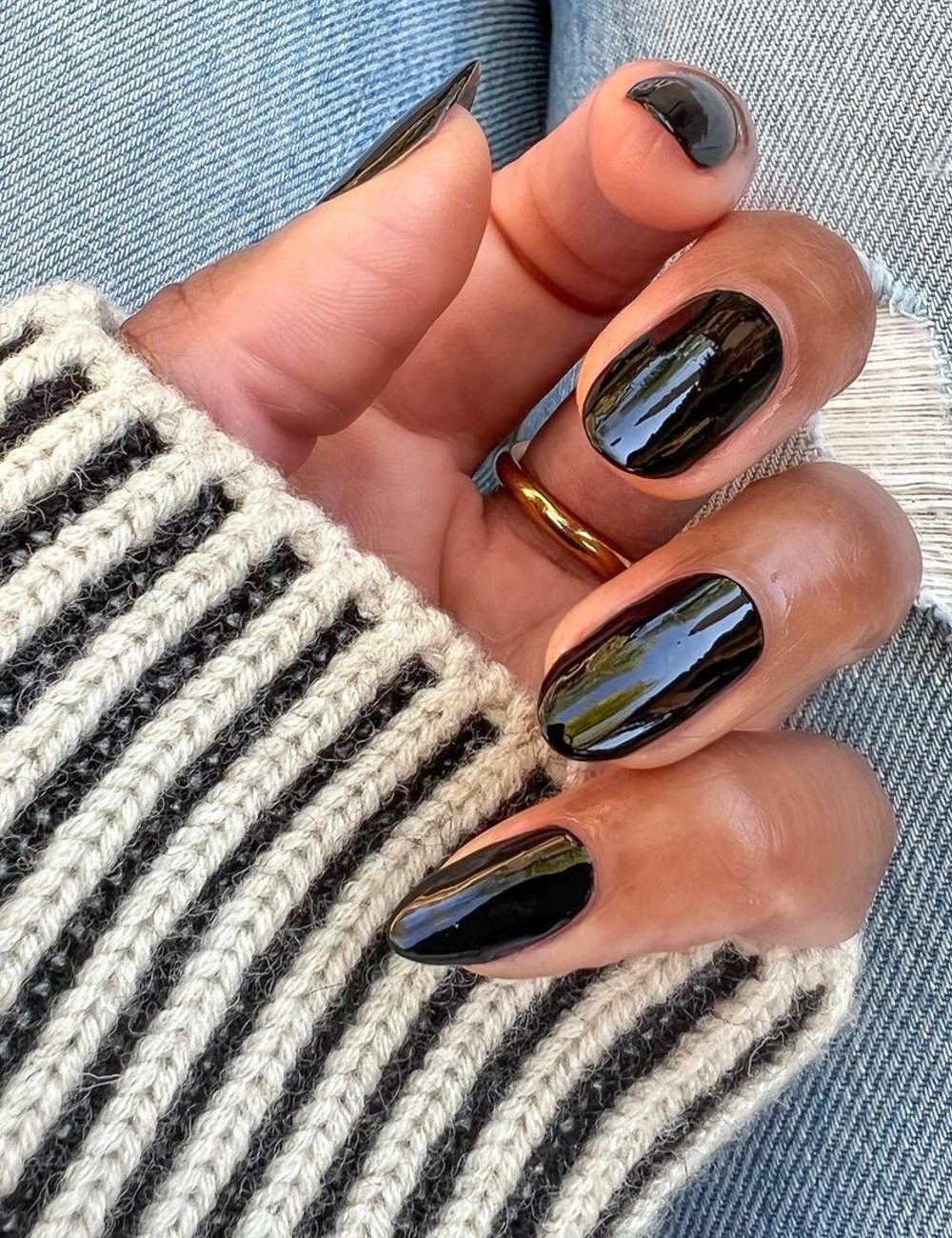 Unhas pintadas com cor de esmalte preto brilhante, tendência de moda atual, em foco. Mão usando um suéter listrado branco e preto, e jeans claro ao fundo, exemplificando um estilo casual e moderno.