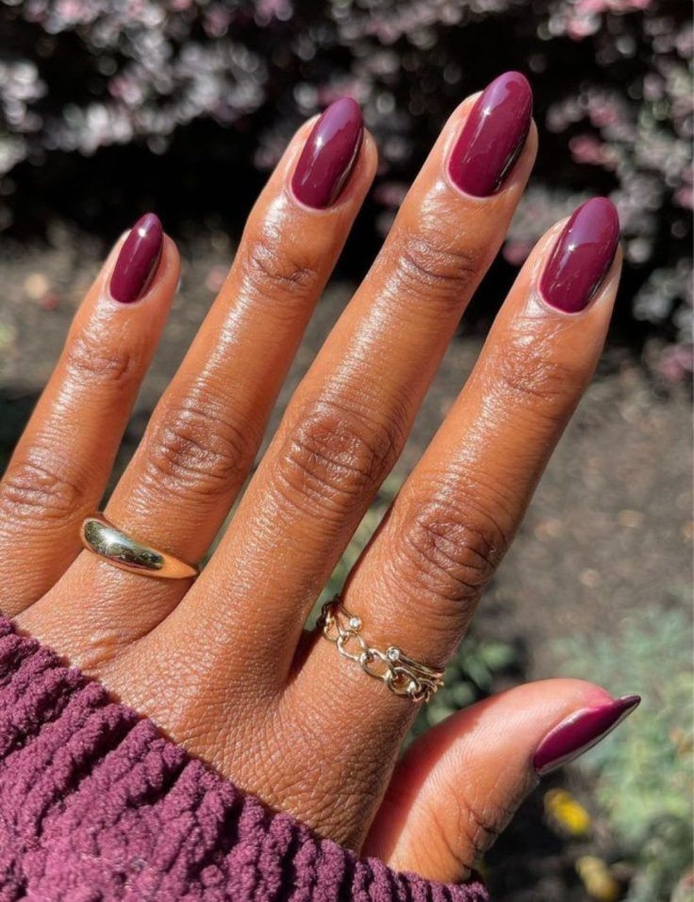 Unhas ovaladas com cor de esmalte roxo, tendência elegante e sofisticada. A mão usa anéis dourados delicados e uma blusa de tricô roxa, destacando um estilo moderno e requintado. Perfeita combinação de acessórios e moda outonal.