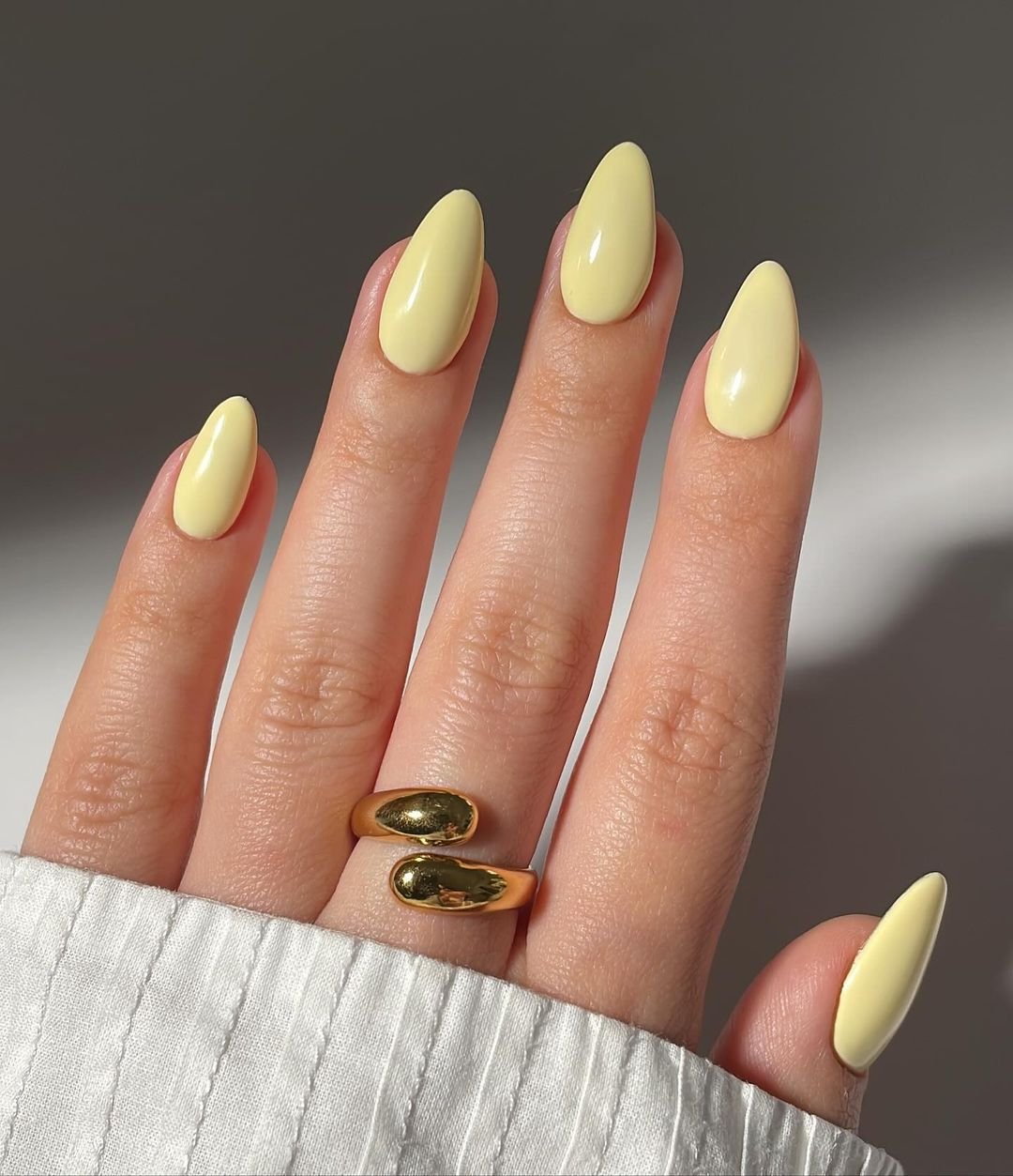 Unhas decoradas com esmalte amarelo pastel em formato amendoado, combinadas com anéis dourados modernos e detalhe de mangas de camisa branca com tecido texturizado. Tendência atual destacando tons suaves e joias minimalistas.