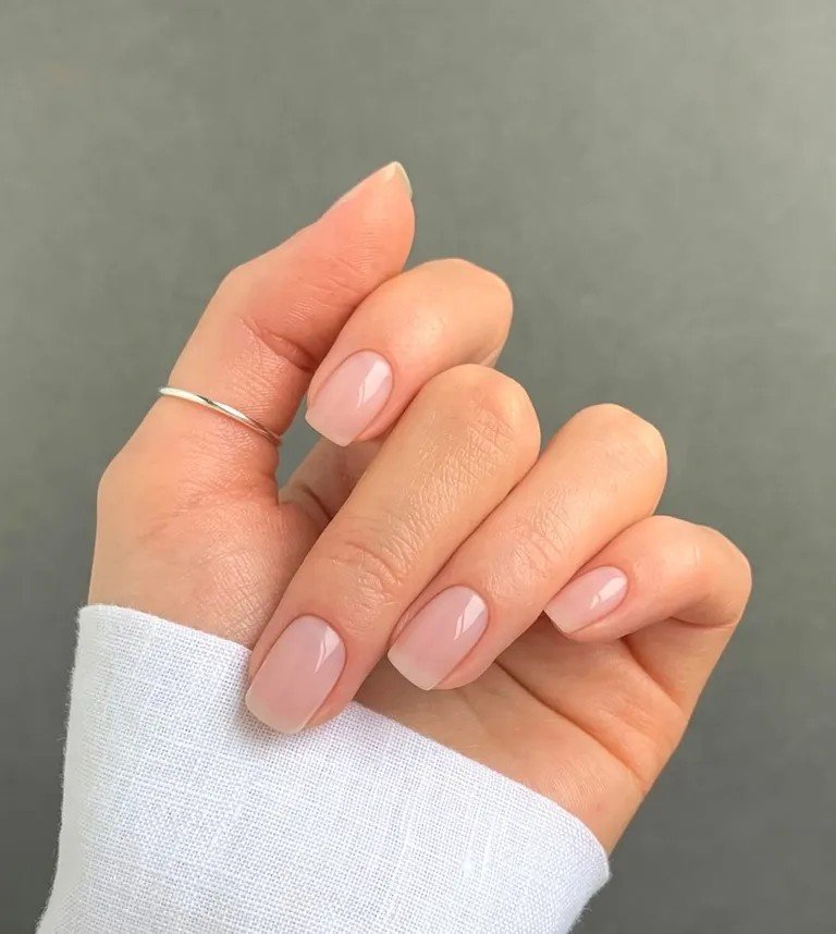 Unha com esmalte nude, acabamento brilhante, em mãos com anel delicado de prata. Manga de blusa branca texturizada cobre parcialmente o pulso. Look minimalista e elegante, destacando tendências de manicure natural e acessórios simples.