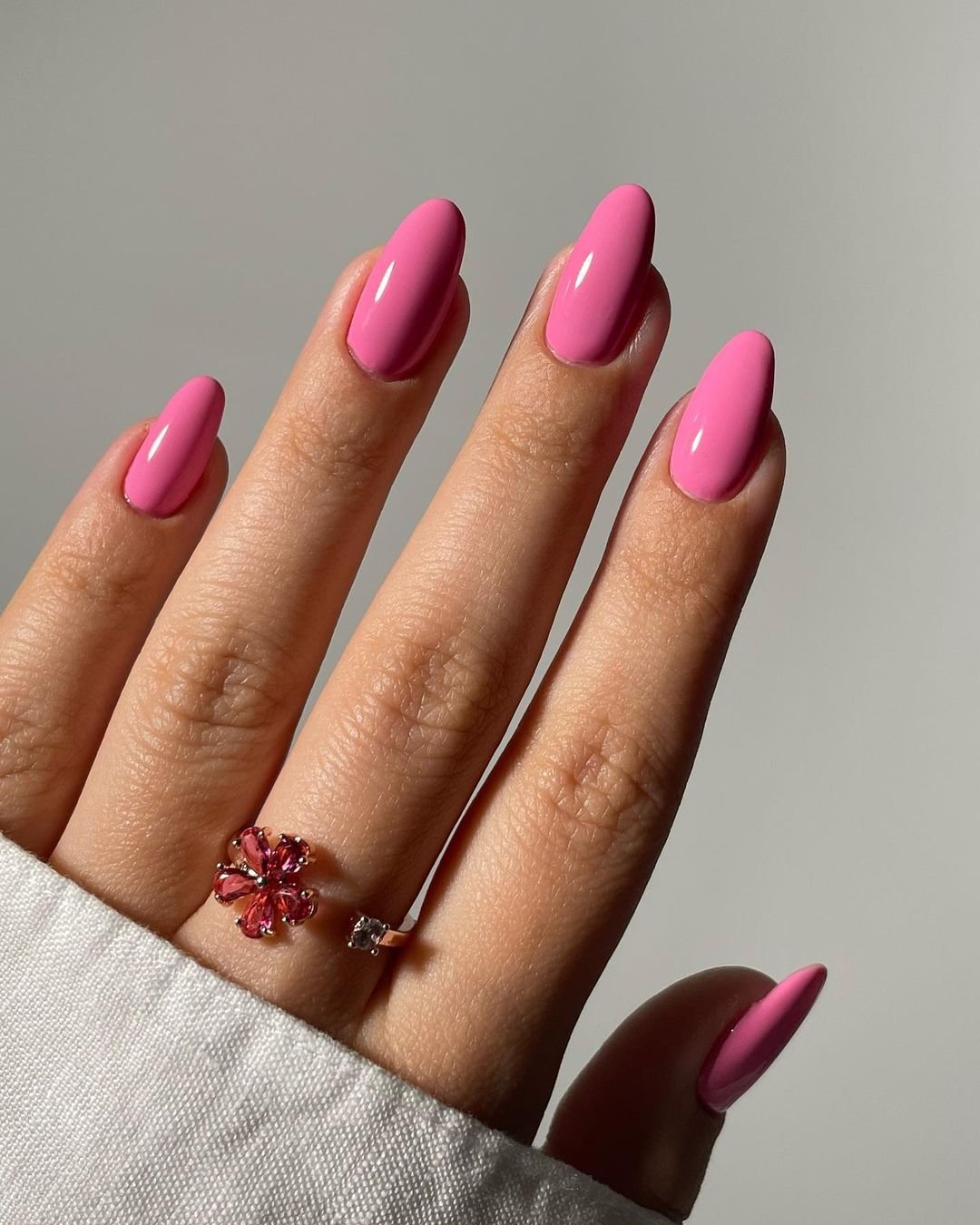 Unhas compridas e ovais decoradas com esmalte rosa vibrante, destacando a tendência de unhas coloridas. Mão usando anel delicado com flor rosa, combinando com a cor das unhas. Moda de acessórios minimalistas e manicure sofisticada.