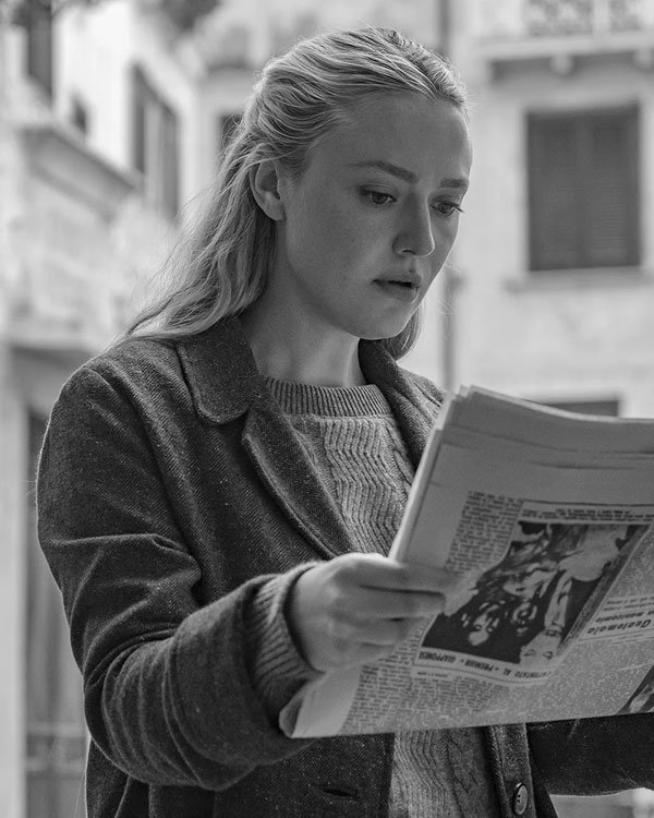 Mulher lendo jornal veste suéter cinza e casaco escuro, combinando moda casual e confortável. O suéter de malha grossa adiciona um toque clássico ao visual, enquanto o casaco transmite elegância prática e atemporal.