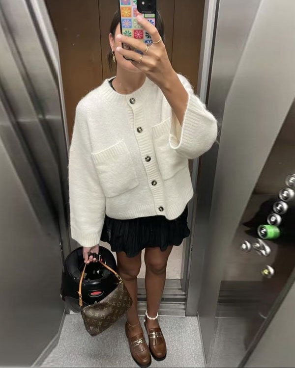 Look outono/inverno com cardigan branco oversized, saia curta preta plissada, sapatos loafer marrom e meias brancas. Acessórios: bolsa Louis Vuitton e outra bolsa preta. Tendências: camadas, conforto e mix de texturas.