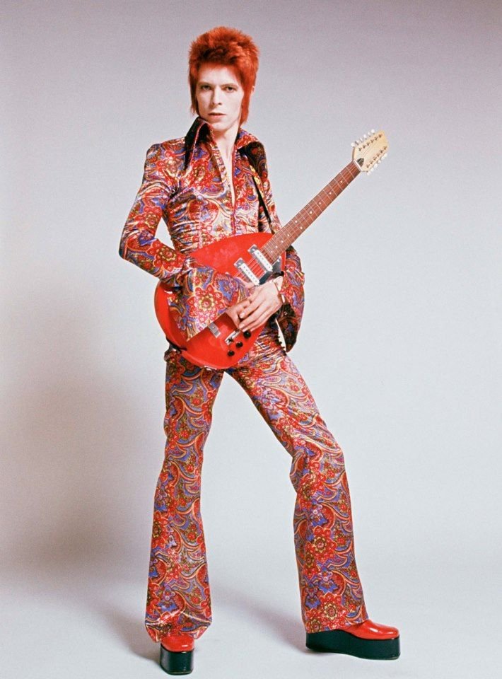 Imagem de uma pessoa com estilo glam rock dos anos 70, vestindo um macacão de estampas psicodélicas coloridas, sapatos plataforma vermelhos e segurando uma guitarra vermelha. A moda destaca extravagância, cores vibrantes e atitude ousada.