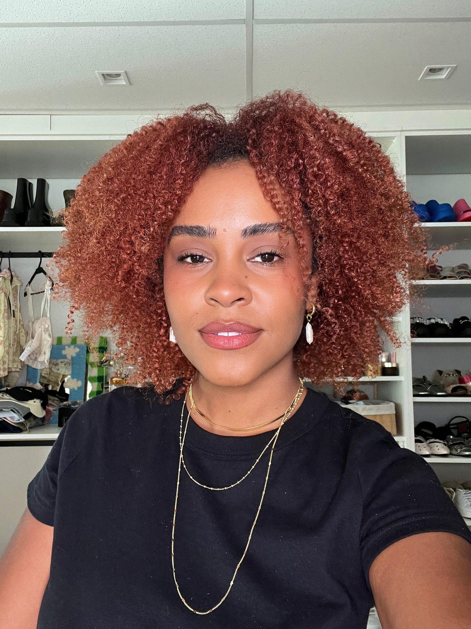 Mulher com cabelo cacheado vermelho, vestindo camiseta preta básica. Acessórios incluem colares dourados em camadas e brincos de concha, refletindo tendências minimalistas e naturais na moda. dicas de maquiagem