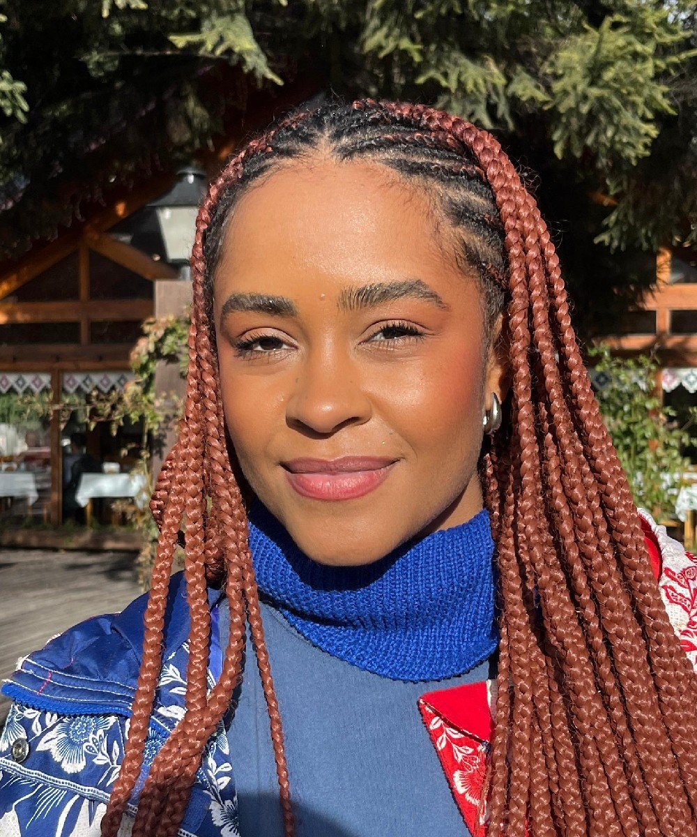 Mulher com tranças longas estilo box braids em cobre, usando suéter azul vibrante e jaqueta estampada com elementos florais, combinando tendências de moda contemporânea e estilo afro. dicas de maquiagem