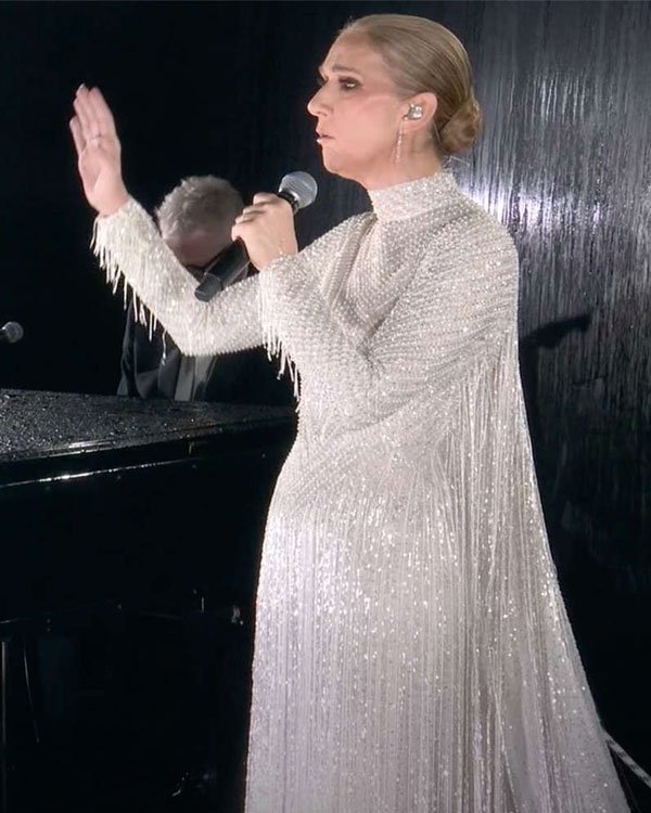 Cantora usando um vestido de gala longo, brilhante e de cor clara, com manga longa e detalhes de franja, em uma apresentação. O visual reflete uma tendência de moda elegante e glamourosa, ideal para eventos formais e performances artísticas.