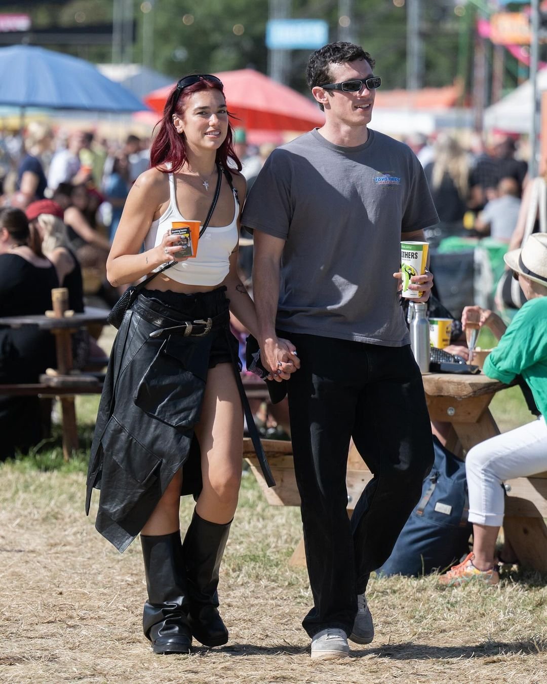 Casal em evento ao ar livre. Ela veste top branco, jaqueta preta amarrada na cintura, botas pretas e óculos de sol. Ele usa camiseta cinza, calça preta e óculos escuros. Estilo casual e descontraído, com influências de moda urbana.