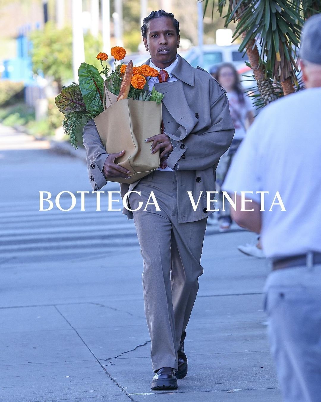 Homem com um estilo elegante, vestindo um terno bege oversized e trench coat da Bottega Veneta. Ele carrega um buquê de flores e baguetes, destacando uma tendência de moda casual e sofisticada, ideal para o ambiente urbano.