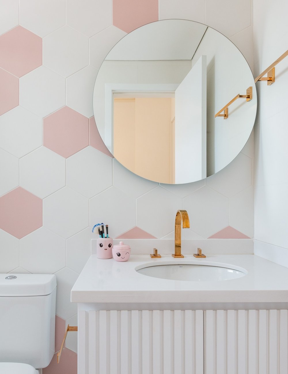 Lavabo moderno com revestimento de hexágonos rosas e brancos, espelhos de banheiro redondo, torneira dourada, bancada branca e acessórios decorativos em tons de rosa. Design minimalista e elegante, seguindo tendências contemporâneas de decoração.