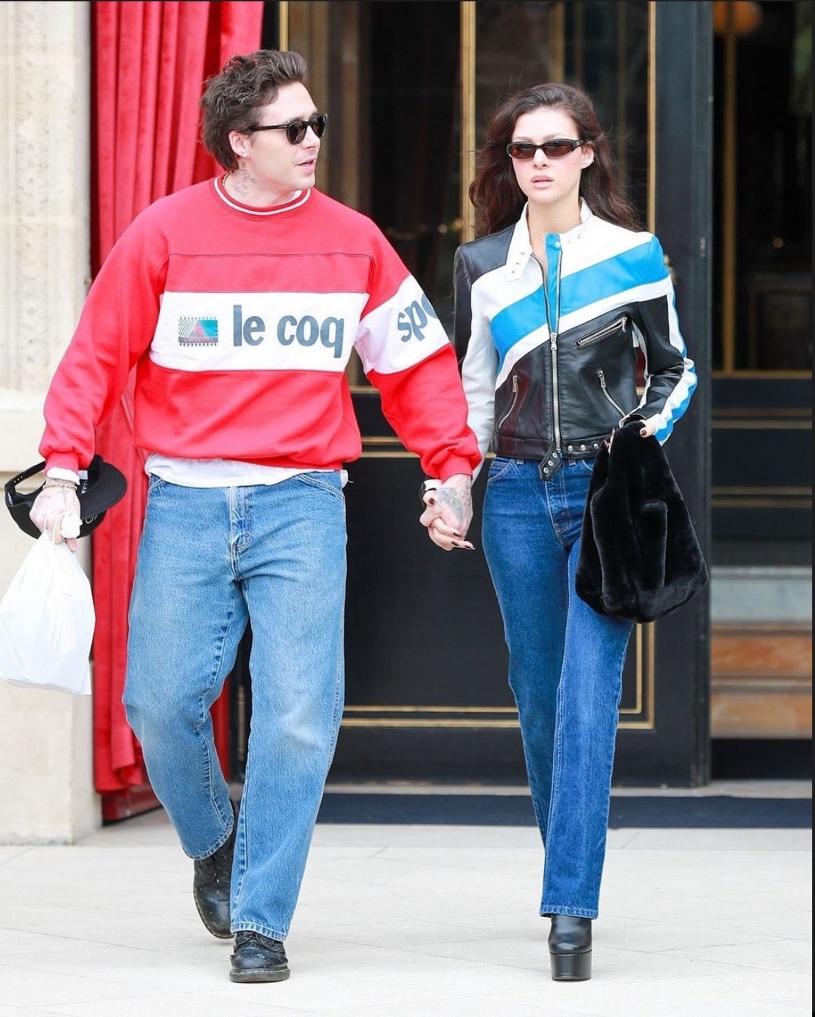 Casal com estilo despojado e moderno. Ele usa moletom vermelho com logo esportivo, calça jeans larga e tênis negros. Ela veste jaqueta de couro bicolor e jeans skinny, combinados com botas plataforma pretas e óculos de sol.