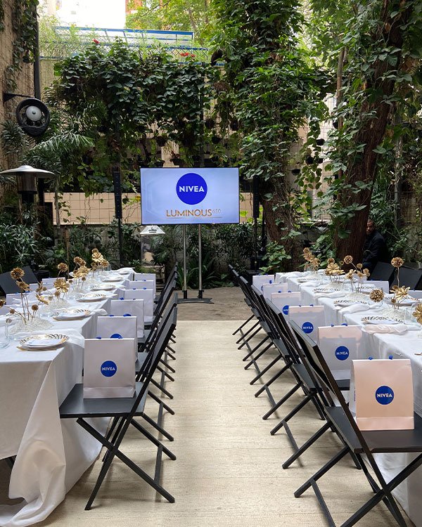 Evento Nivea Luminous 630 em ambiente ao ar livre cercado por vegetação. Decoração elegante com mesas cobertas por toalhas brancas, cadeiras pretas dobráveis e sacolas de presentes Nivea nos assentos. Tendências clean e minimalistas em destaque.