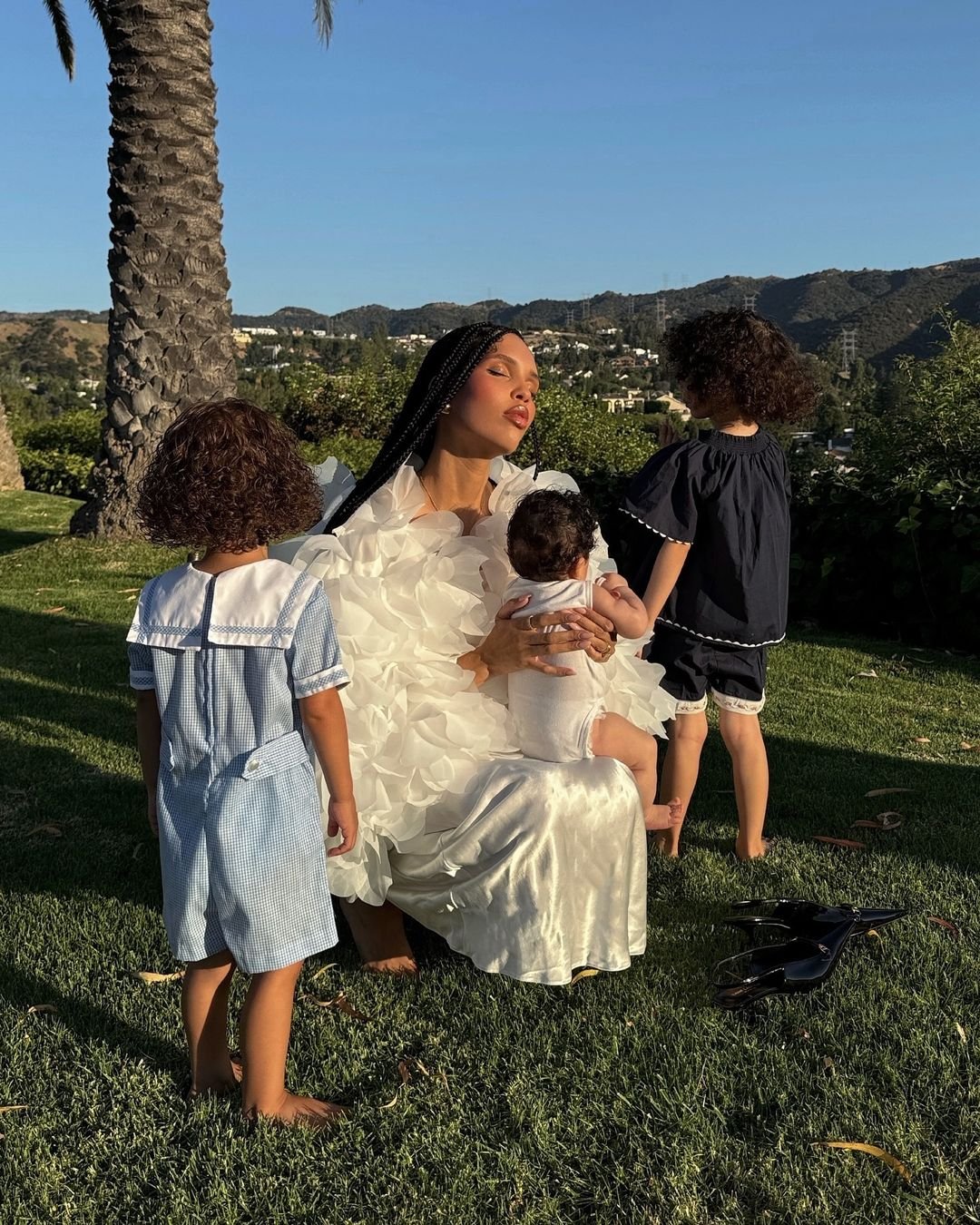 Imagem de mulher usando vestido branco volumoso e sapatos pretos ao lado, acompanhada por três crianças. As crianças vestem roupas infantis de estilo clássico em tons de azul claro e marinho. Fundo com paisagem de montanhas e árvore. baby blues