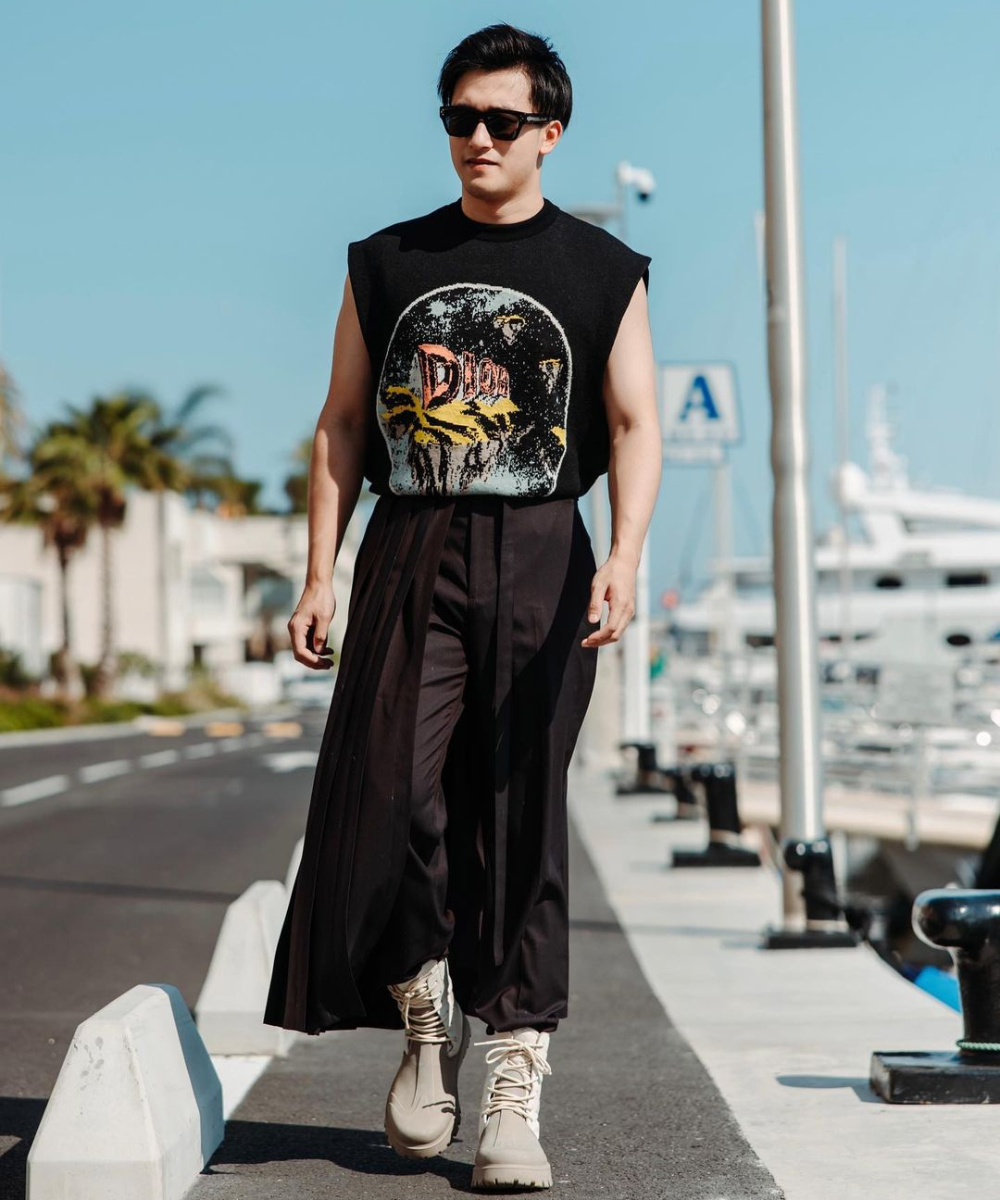 Homem vestindo regata preta com estampa de banda, calça larga preta estilo samurai, botas bege, óculos de sol escuros. Look urbano e moderno, mistura de estilos casuais e alternativos. Cenário de área portuária ao fundo.