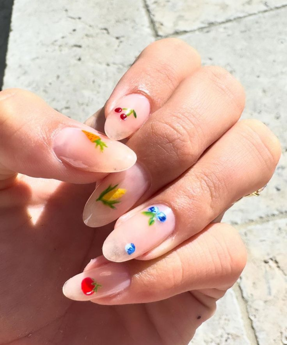 Unhas decoradas com estilo minimalista, apresentando desenhos delicados de frutas e flores sobre uma base transparente. Tendência popular em nail art, destacando-se pela elegância e sutileza, ideal para quem busca um visual moderno e discreto.