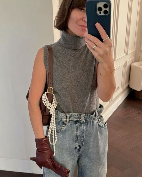 Pessoa usa uma blusa cinza sem mangas, calça jeans de cintura alta, luvas marrons e uma bolsa de ombro com detalhes de pérolas. Moda casual chic, mistura de elementos clássicos com toque moderno.