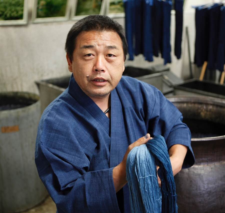 Homem vestindo um traje tradicional japonês indigo, kimono, destacando-se na tradição da tinturaria. Ambiente indicativo de um ateliê de tingimento artesanal, com tonéis ao fundo.