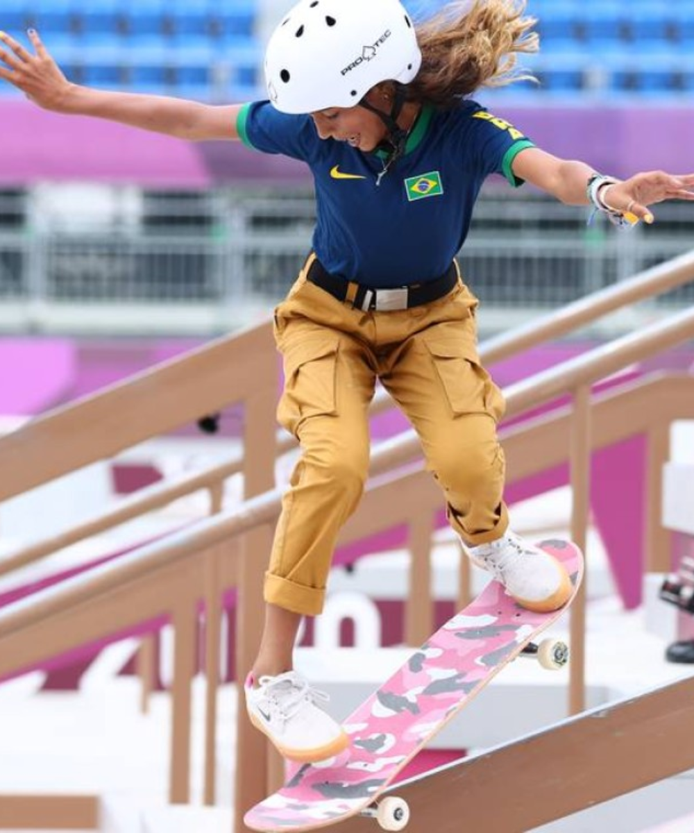 Atleta de skate vestindo uniforme esportivo com camisa azul, bandeira do Brasil, calça cargo khaki e tênis branco. Capacete branco para proteção e skate com estampa camuflada rosa. Moda casual e prática, destacando tendências esportivas urbanas.