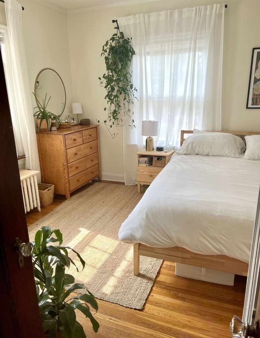 Quarto decorado em estilo minimalista, com móveis de madeira clara e plantas. Cortinas brancas permitem a entrada de luz natural, criando um ambiente iluminado e acolhedor. Ideias para quarto pequeno.