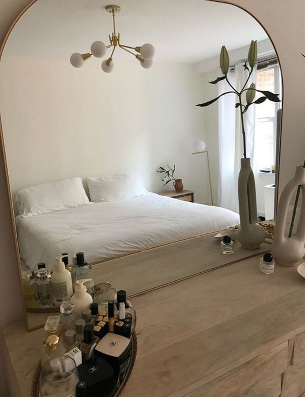 Decoração de quarto minimalista, com paleta de cores neutras. Destaque para espelho grande com moldura dourada, cama com roupa branca, luminária moderna e arranjos de flores em vasos. Ideias para quarto pequeno