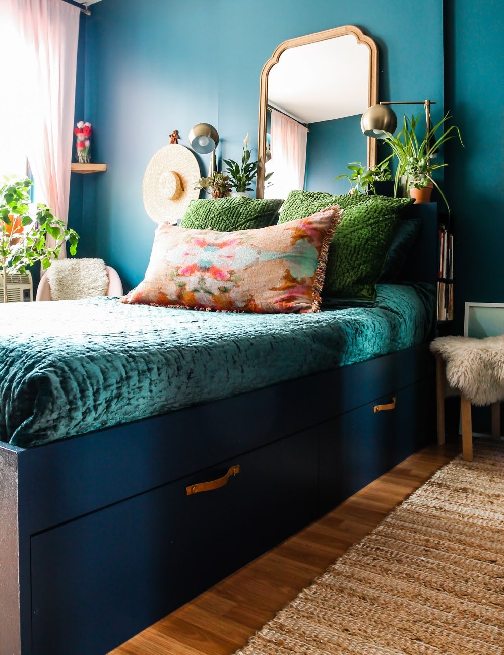 Quarto moderno com parede azul, cama de veludo verde, almofadas coloridas e decoração elegante. Tendências de decoração incluem plantas, espelhos ornamentados e iluminação suave, criando um ambiente aconchegante e estiloso. Ideias para quarto pequeno