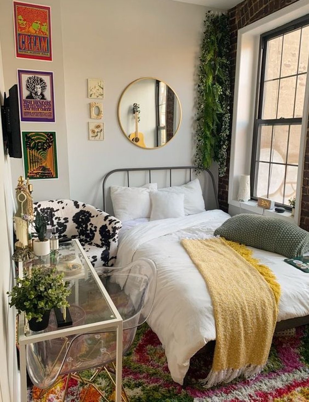 Quarto decorado com estilo boho chic, apresentando móveis modernos e plantas como destaque. Cadeira transparente, pôsteres vintage na parede, cadeira de pelúcia preta e branca, cama com edredom branco e manta amarela. Ideias para quarto pequeno.