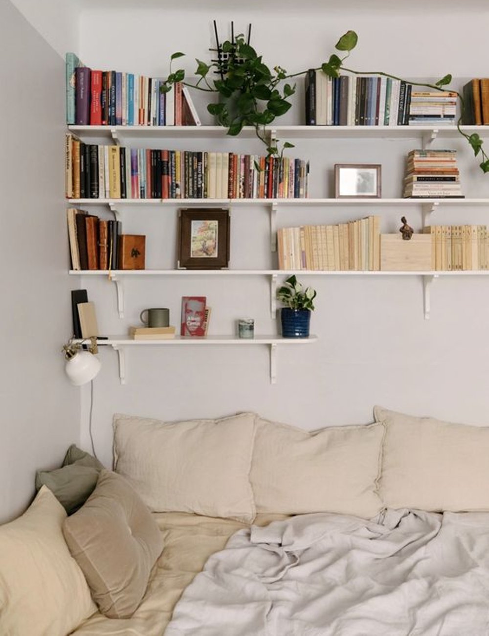 Quarto aconchegante com cama de tons neutros e almofadas macias. Prateleiras abertas na parede exibem uma coleção de livros e plantas, realçando uma decoração minimalista e moderna com um toque natural e rústico. Ideias para quarto pequeno.