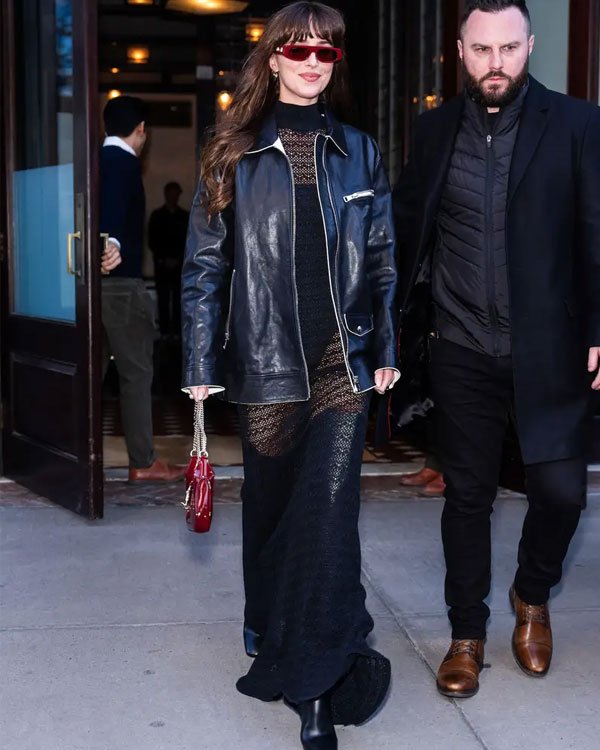 Mulher usando casaco de couro preto, vestido longo preto semi-transparente, óculos de sol vermelhos e carregando uma bolsa vermelha. Moda urbana com toques de elegância e ousadia, combinando peças clássicas e modernas.