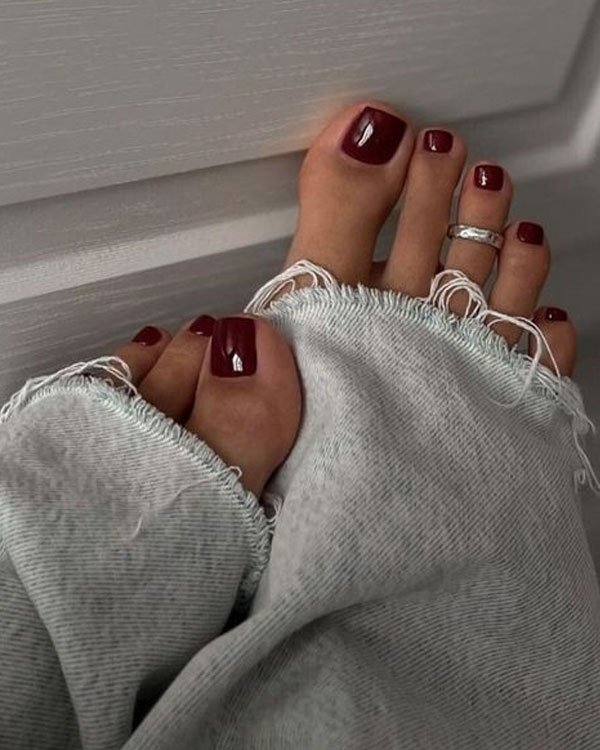 Imagem de pés com unhas pintadas de vermelho escuro, usando um anel no dedo do meio. A calça jeans desgastada na barra revela a tendência casual. A moda destaca detalhes rústicos e acessórios minimalistas, combinando conforto e estilo.