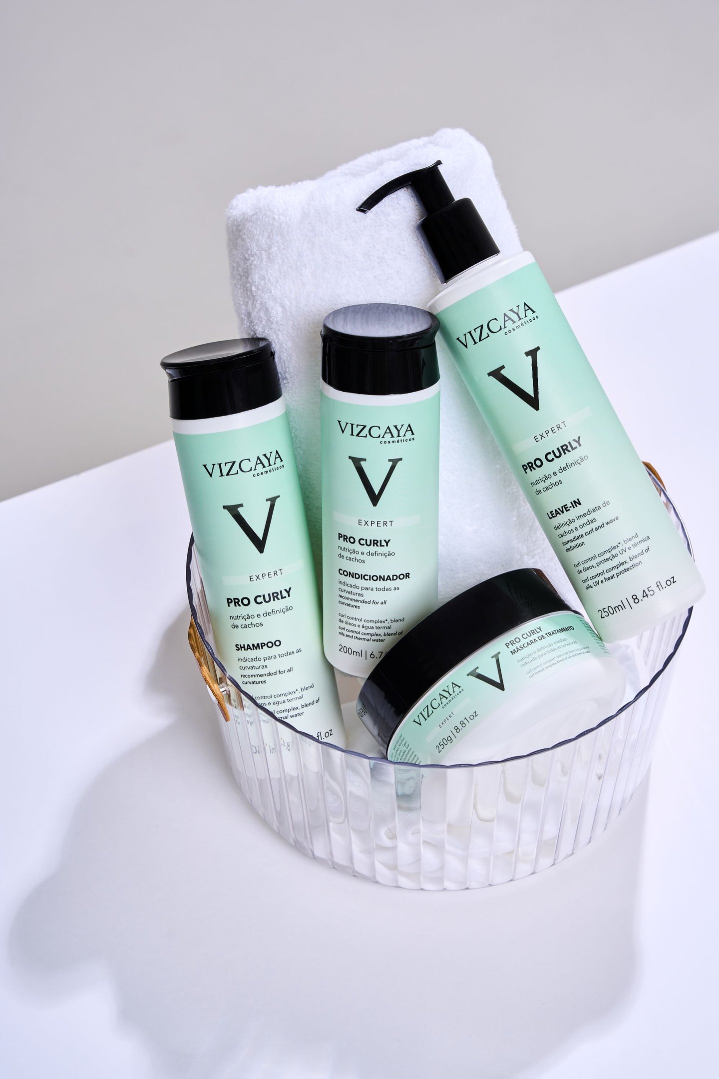 Vizcaya Pro Curly para cabelos cacheados, incluindo shampoo, condicionador, leave-in e máscara de tratamento, em um recipiente transparente ao lado de uma toalha branca. Embalagens modernas em verde claro com detalhes preto. lançamentos de beleza