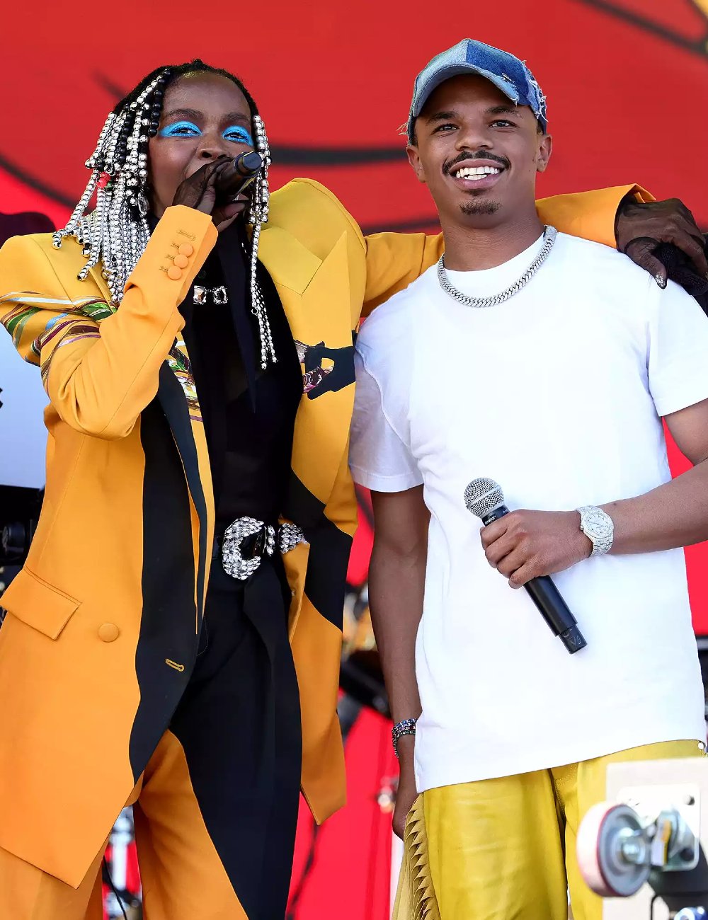 Lauryn Hill vestindo terno amarelo e preto com acessórios, incluindo miçangas no cabelo e maquiagem destacada. Ao lado dela, YG Marley com camiseta branca e calça dourada, usando boné jeans e corrente prateada.