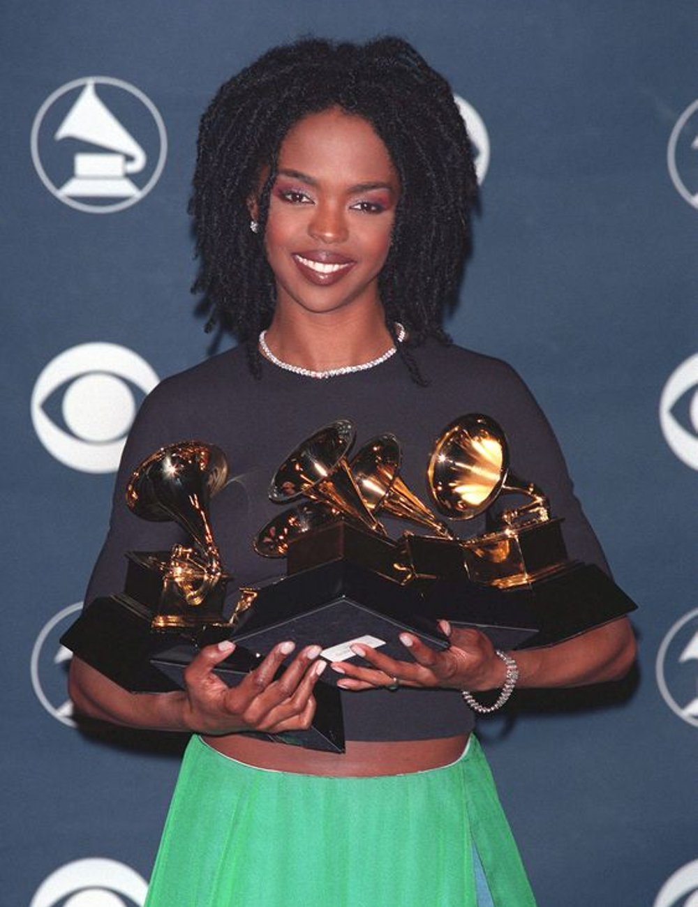 Lauryn Hill segura diversos prêmios usando roupa marcante: cropped preto e saia verde. Look é complementado com colar de pérolas e cabelos no estilo dreadlocks, evidenciando uma combinação de elegância e estilo casual.