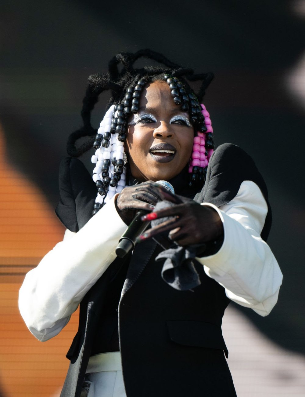 Lauryn Hill com cabelo estilizado com miçangas preto, branco e rosa, maquiagem artística destacada em branco prateado, vestindo um terninho preto com detalhes em branco, destacando ombreiras amplas.