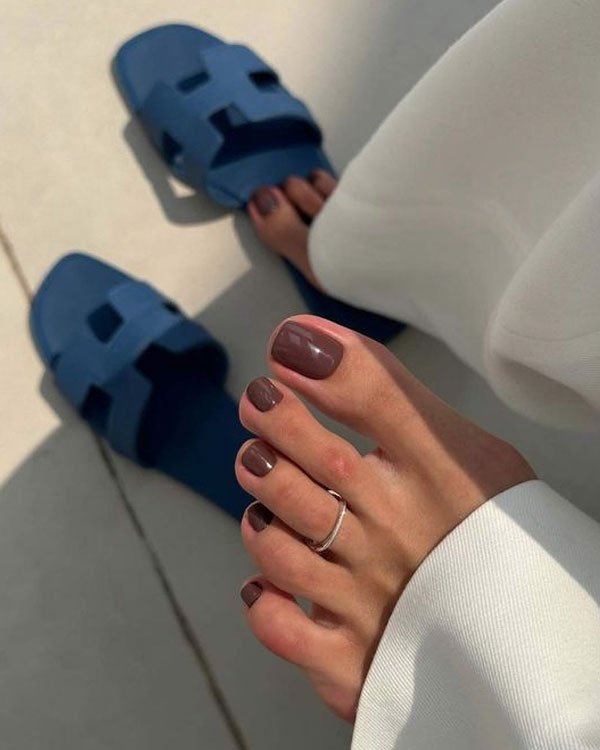 Sandálias azuis estilo slide, unhas dos pés esmaltadas em tom marrom com um anel delicado no dedo. Calça branca casual. Estilo minimalista e elegante, perfeito para dias de verão. Tendência: acessórios discretos e conforto.