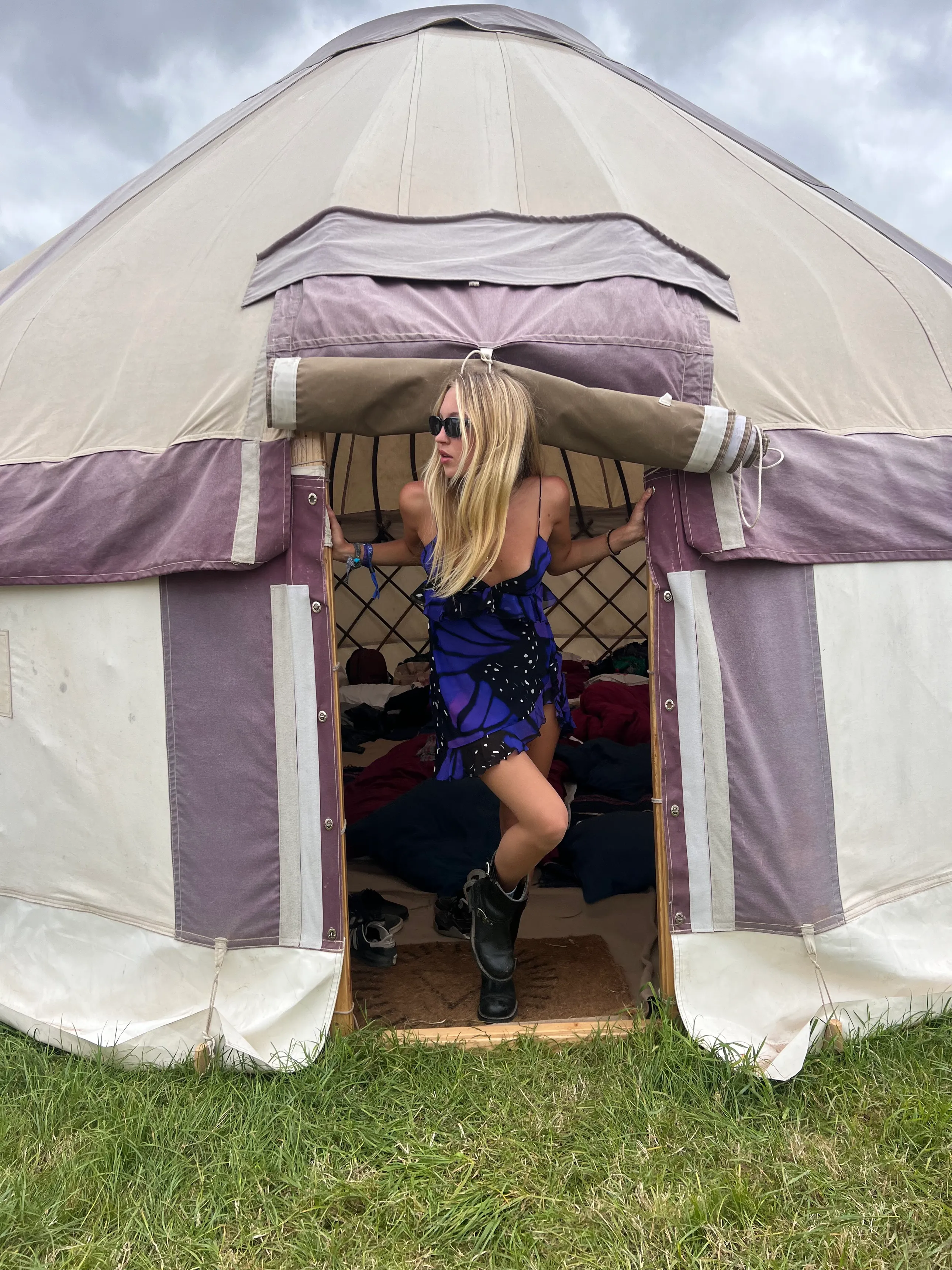 Mulher com vestido curto roxo e preto, óculos escuros, e botas pretas, saindo de uma tenda estilo yurt. Visual moderno e descontraído, evidenciando influência boho e fashion de festivais. Fundo de gramado e céu nublado.