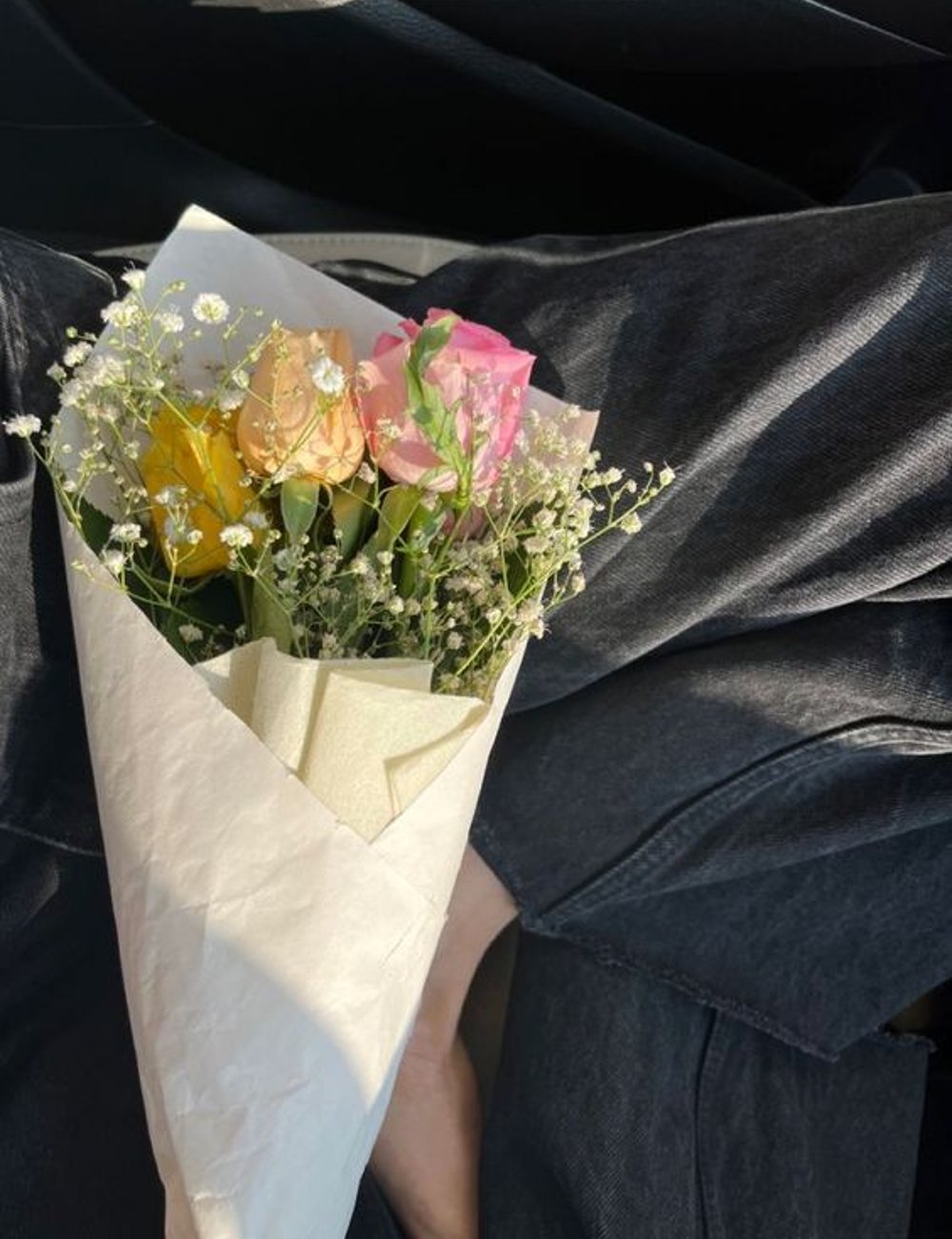 Close-up de um buquê de flores com rosas amarelas, rosas e brancas em fundo de jeans preto. Moda casual com calças jeans de lavagem escura, tendência atual de looks minimalistas e confortáveis. Linguagem do amor.