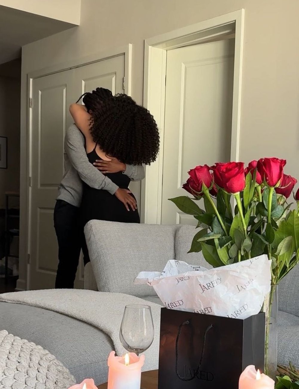 Casal abraçado em uma sala de estar bem decorada. Ela veste um vestido preto justo e ele uma camiseta cinza e calça preta. Decoração romântica com velas acesas e buquê de rosas vermelhas. Linguagem do amor.