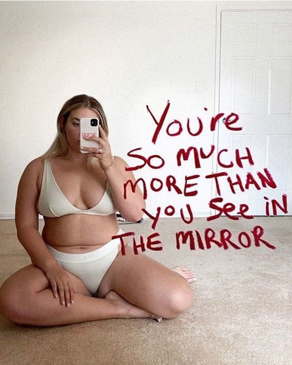 Mulher plus size veste lingerie bege de dois peças e tira selfie no espelho. A imagem contém mensagem de autoaceitação escrita no espelho. Tendência em moda íntima confortável e inclusiva, celebrando a diversidade de corpos.