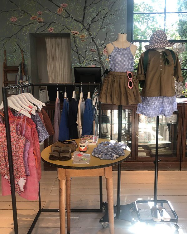 Vitrine exibindo roupas infantis em tendência, como vestidos florais, bermudas coloridas, conjuntos de tecidos leves e sandálias em tecido. Mix de estampas e cores vibrantes é destaque, seguindo a moda casual e confortável para crianças.