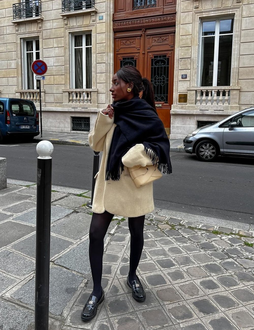 Pessoa vestindo look para frio simples: sobretudo bege, meia-calça preta e sapatos estilo loafers. Usa cachecol preto, bolsa pequena e brincos dourados. Look elegante e urbano.