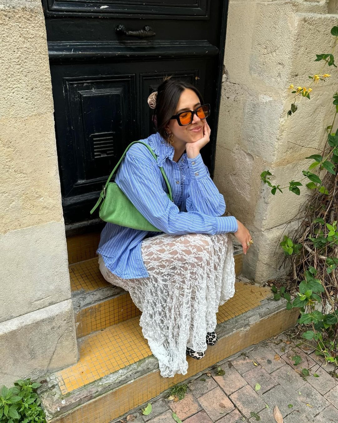 Mulher sentada em degrau vestindo camisa azul listrada oversized, saia longa de renda branca, óculos de sol laranja, bolsa verde e laço no cabelo. Look combina estilos casual e romântico, destacando tendências como camadas de renda e acessórios coloridos.