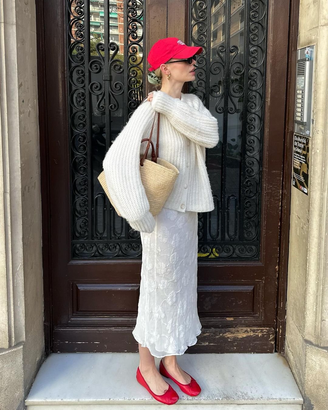 Pessoa vestindo um suéter de tricô branco oversized, saia midi branca texturizada, sapatilhas vermelhas e boné vermelho. Carrega uma bolsa de palha. Estilo casual chique com combinação de tons neutros e acessórios vibrantes. Tendência: contraste de cores.