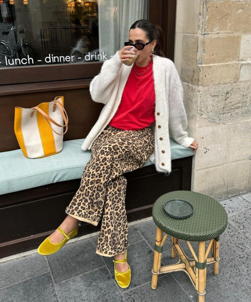 Mulher com estilo urbano eclético: cardigan de lã branco, camiseta vermelha, calça com estampa de oncinha e sapatilhas amarelas de veludo. Completa o look com bolsa bicolor e óculos escuros. Tendência mistura de estampas e texturas.