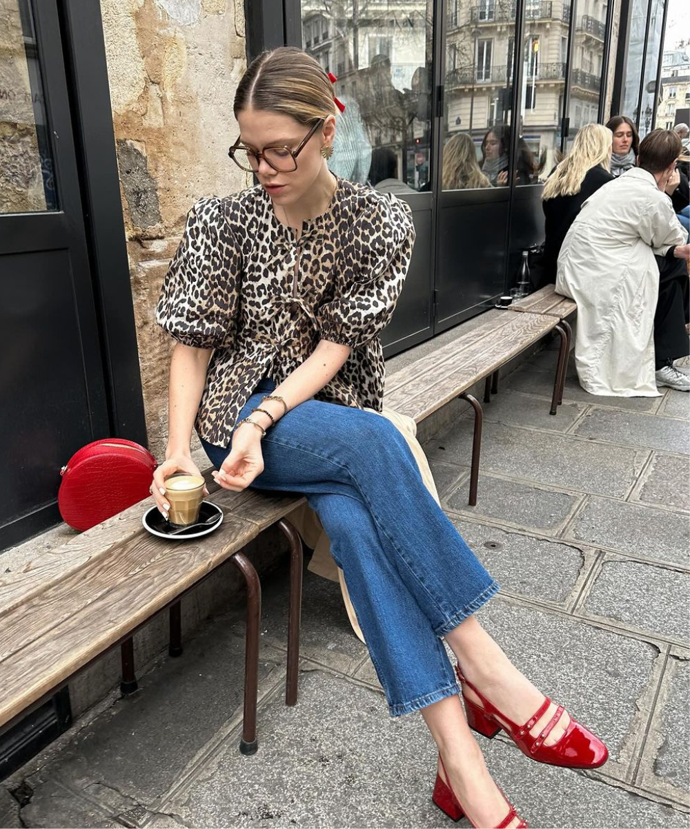 Mulher usando blusa de estampa animal print, calça jeans reta e sapatilhas vermelhas brilhantes. Acessórios incluem óculos de armação grossa, brincos dourados e bolsa vermelha pequena. Look casual sofisticado e moderno, popular em tendências urbanas.